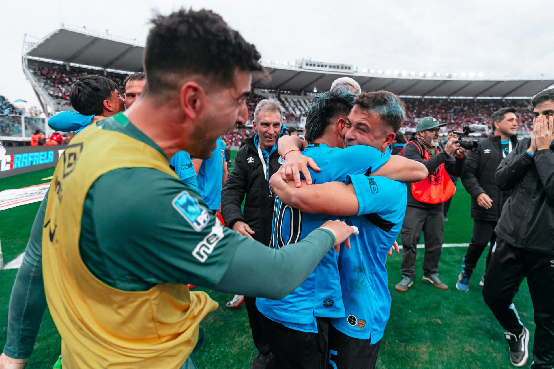 Compañero corriendo hacia el abrazo de dos jugadores de Belgrano